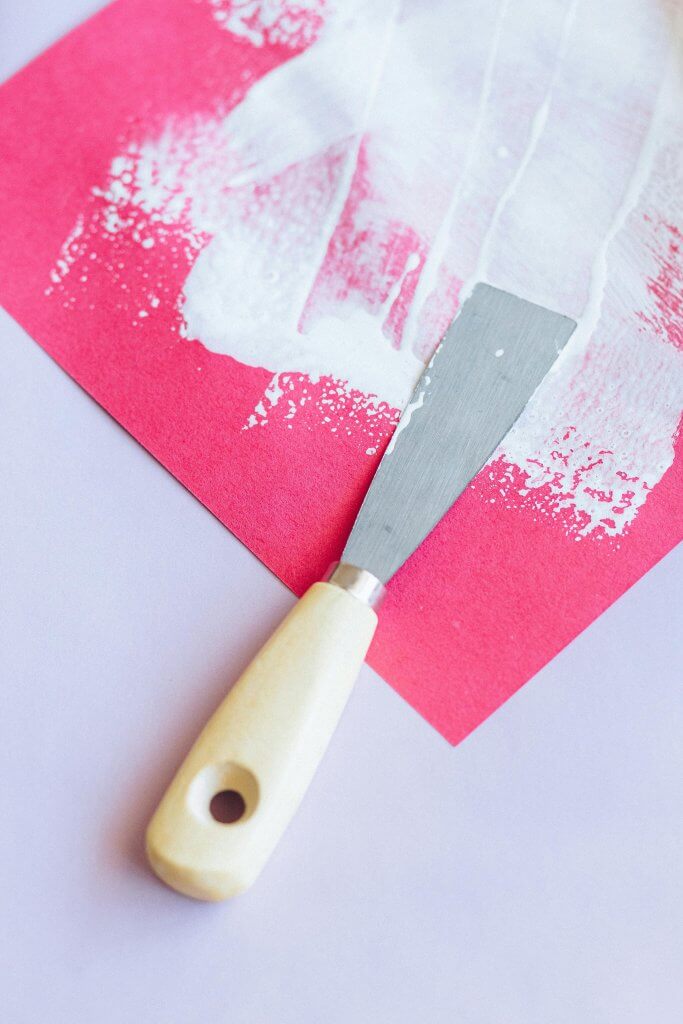 Image avec une spatule de peinture sur une feuille de papier rose avec de la peinture blanche